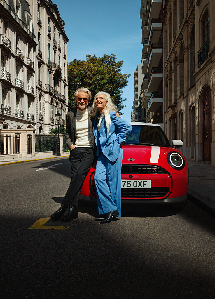 Zwei Personen stehen vor einem roten MINI Cooper mit weißem Dach an einer städtischen Straße.